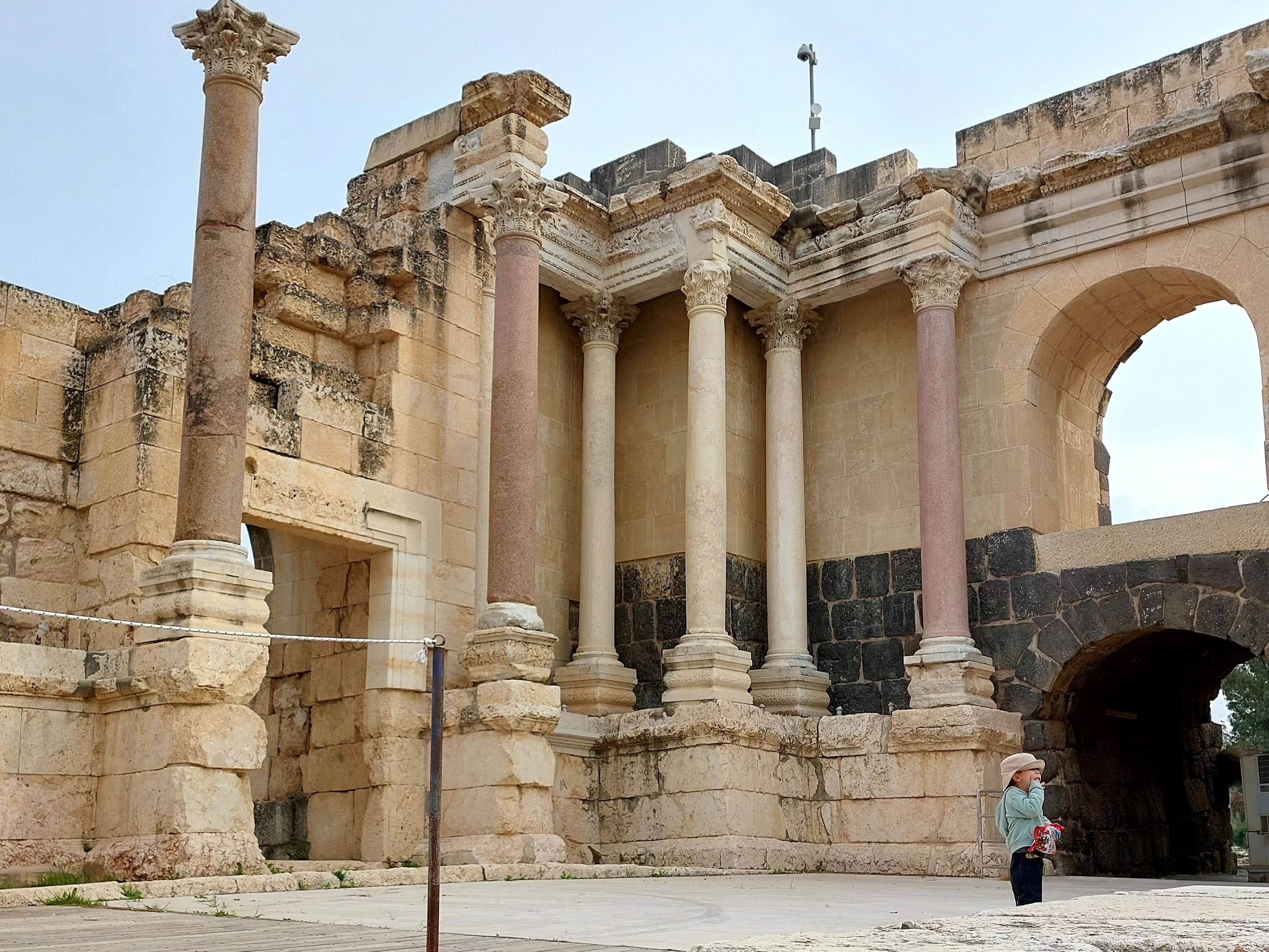 Close-up detail of the Roman theater at Beth-Shan showing ornate Corinthian columns with pink granite shafts against limestone stonework, with a small child standing at the base for scale