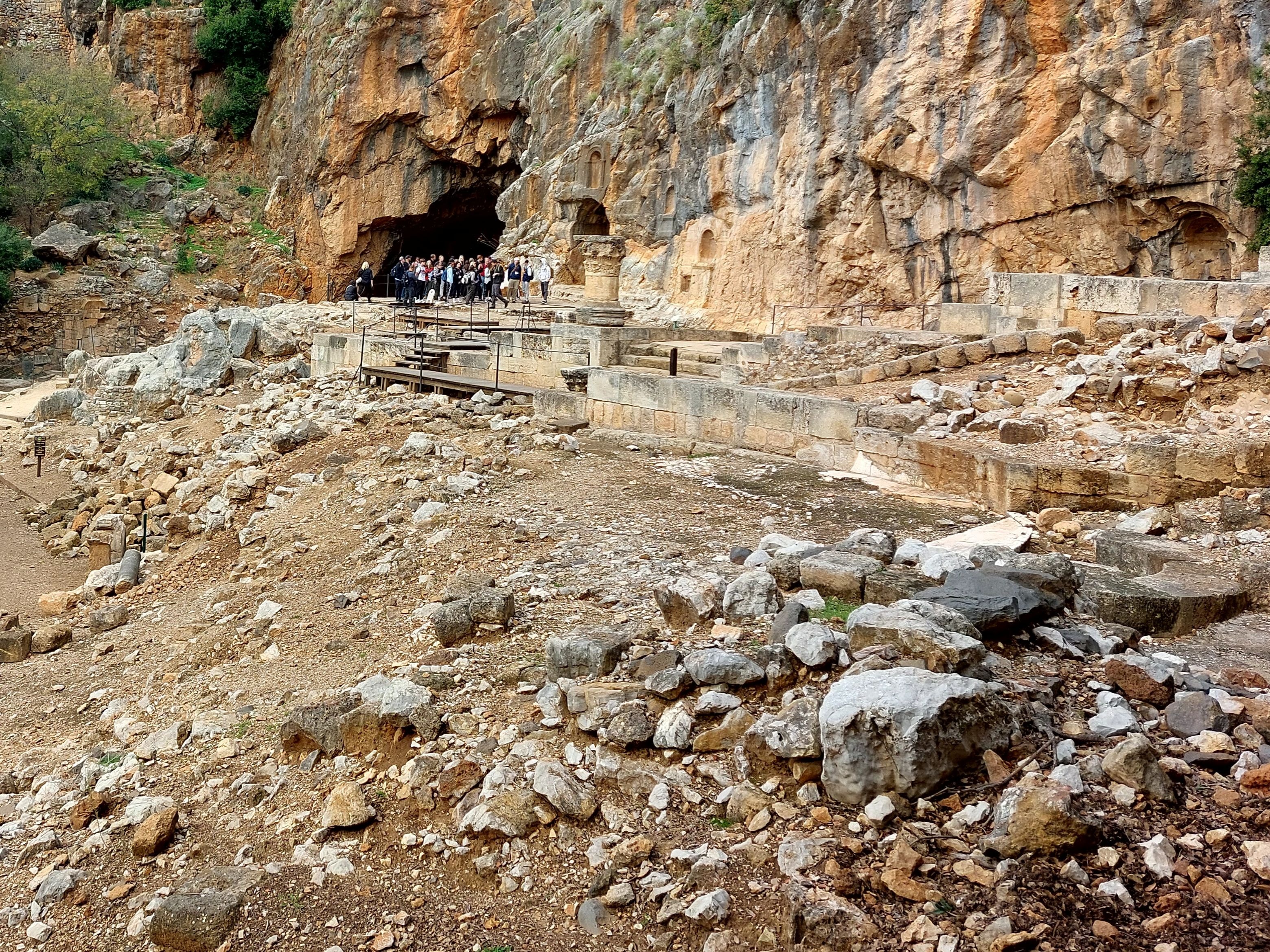 Wide view of the Caesarea Philippi ruins with scattered boulders and temple foundations in the foreground and a tour group gathered at the entrance of the cave of Pan carved into the dramatic cliff face behind them