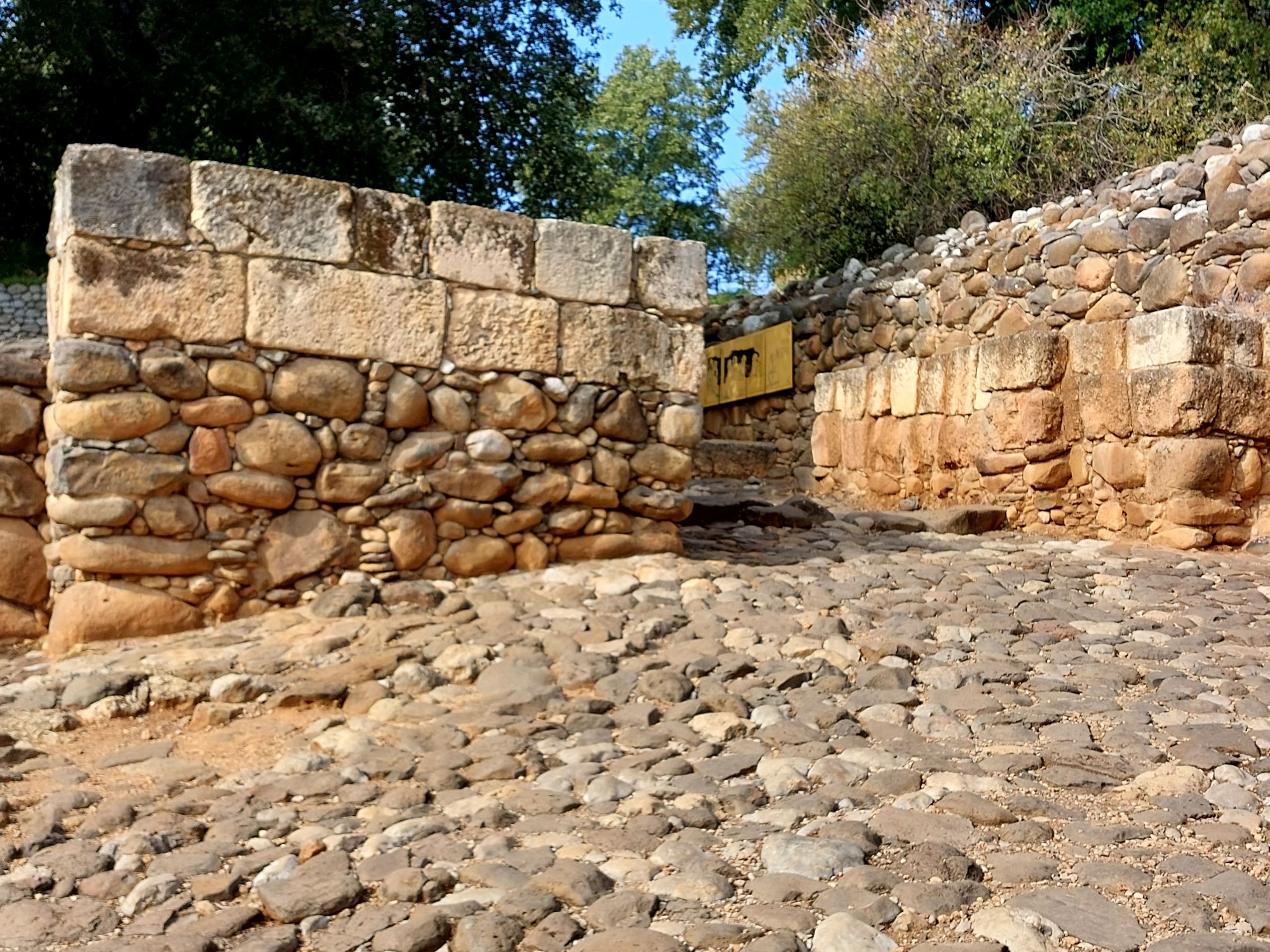 Ancient cobblestone road leading through the gap between massive stone walls at the gate of the city of Dan, with mixed fieldstone and ashlar construction and trees framing the blue sky above