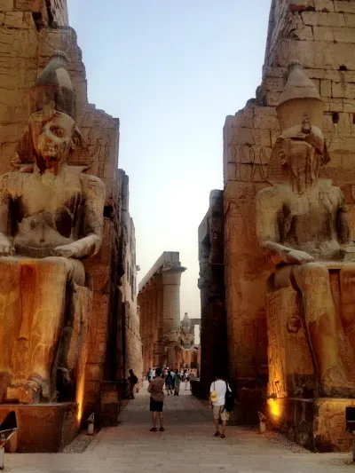 Colossal seated statues of Ramesses II flanking the entrance to Luxor Temple at dusk — visitors walk between the towering ancient Egyptian pharaoh figures