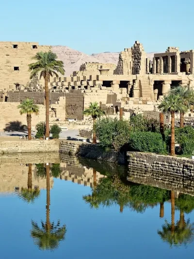 Ancient Egyptian temple ruins reflected in a sacred lake, with palm trees and stone pylons — the ruins of Karnak Temple complex near Luxor
